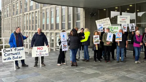 A row of people protesting the plans. They are holding signs. One says 'don't flood cornwall with solar. Another says 'grazing not glasing'