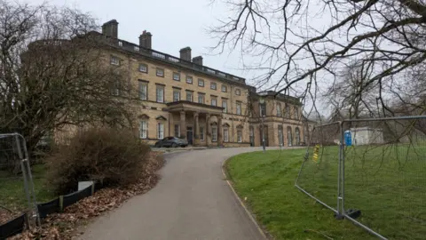 An 18th-century country house, pictured from the driveway leading up to its entrance. Metal fencing and leafless trees can be seen in the foreground.