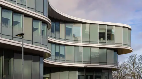 A close-up of a three-storey building with a dramatic glass frontage in the series of curves. Bare trees can be seen in the background on the right.