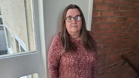 Pearl is standing in the stairwell of a block of flats looking to camera with an unhappy face. She has long brown hair and is wearing black glasses and a red dress with white floral pattern