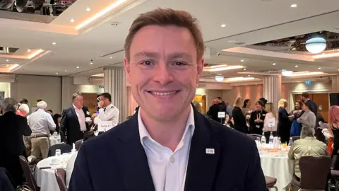 Will Quince stands in a busy conference room filled with round tables, attendees networking, and warm overhead lighting. People are gathered in groups across the room.