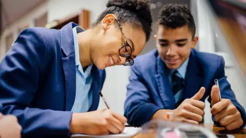Buckinghamshire Mind A girl in the foreground is writing on a pad and smiling while looking at the pad. She wears black-rimmed glasses with tortoiseshell sides and a royal blue school uniform blazer with a light blue shirt. A boy next to her smiles at what she is writing. He wears the same uniform but has a dark blue tie.