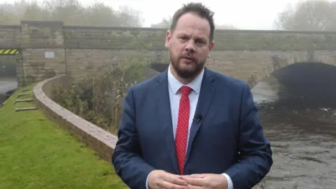 LDRS Simon Lightwood standing outdoors near a stone bridge that spans over a flowing river on a misty day. He is wearing a dark blue suit jacket, a white dress shirt, and a red tie with small white polka dots. A small lapel microphone is clipped to the jacket.
