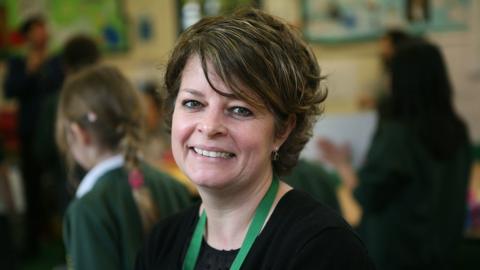 Ruth Perry. She has medium length brown hair, and is wearing a green lanyard around her neck. Out of focus in the background is a classroom full of children.