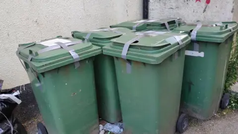 BBC Five green bins in an alleyway, taped up with grey tape