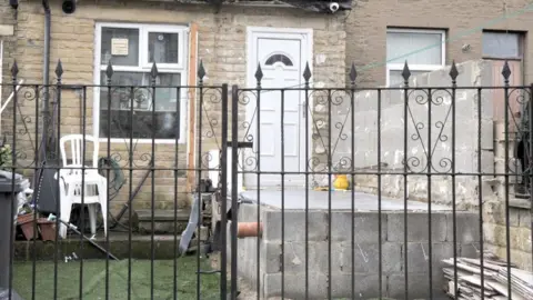 LDRS The photograph shows the ongoing work to remove the extension. The door is now visible through a metal fence, with a concrete structure and a side wall remaining.