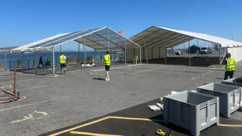 The car park space has two large marquee structures being built, one has a white covering over its roof and two sides. Three people in hi-vis jackets work on the site. You can see the sea in the distance.