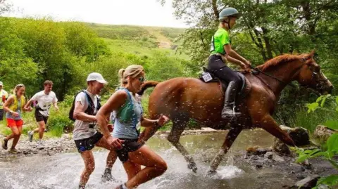 Peter Barnett Two women running next to a horse through a small stream surrounded by fields and treea