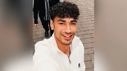 A selfie of a smiling man walking along a pavement wearing a white quarter zip 