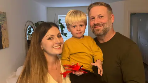 BBC Monika, Henry and Leo smile at the camera in a living room. Monika, who has long brown hair, and Henry who has blonde short hair and a brown jumper are holding toddler Leo who is holding a red toy plane and has a yellow jumper on. 