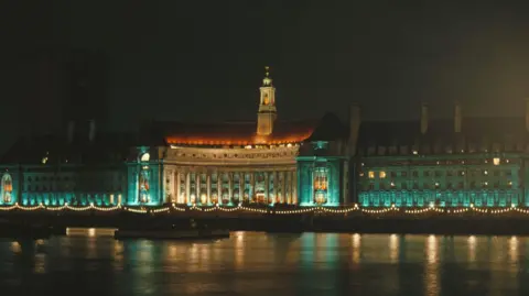 Getty Images The County Hall building on the side of the Thames, viewed from the other side of the river at night. The building is lit up in green, yellow and orange lights.
