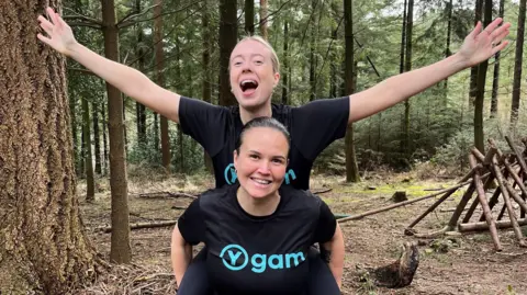 Sisters Michelle Kelly and Emma dressed in running clothing. They are in a forest, with one carrying the other on their back and arms stretched wide. Both are smiling at the camera. 