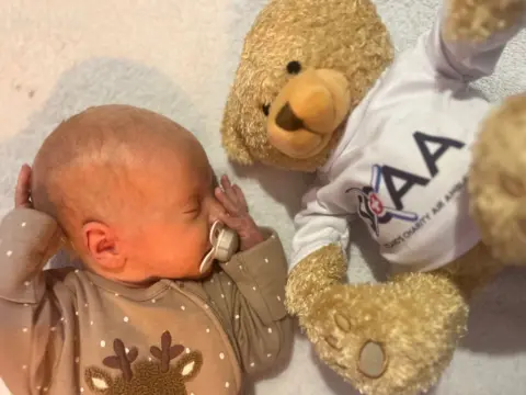Scotland's Charity Air Ambulance/SCAA A baby with a brown grow and a white dummy lies on a white fluffy mat next to a golden teddy bear in a white top, which reads SCAA - Scotland's Charity Air Ambulance.