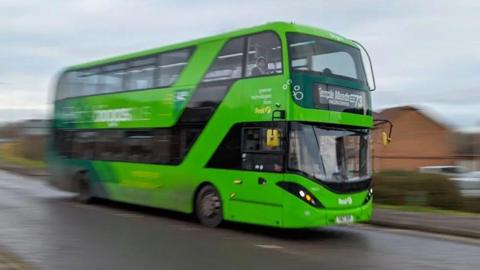 A green double-decker bus in motion. The front of the bus is in focus, while the surroundings are blurred.