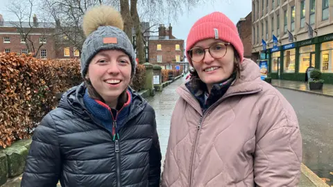 Two women standing on a street - one in a navy coat and hat with a bobble and one in a pink coat and hat with glasses