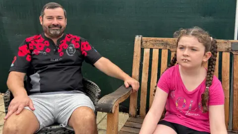 On the left of the image is a a man with short hair and a trimmed beard sitting in a garden chair wearing a black rugby top with red poppies on the shoulders, sitting next to him is his daughter. She has long brown hair in two plaids, wearing a bright pink t-shirt while sitting on a garden bench.