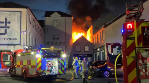 We Are Tyrone Firemen rush to a burning building. Five officers and two vehicles can be seen. 