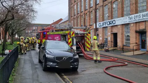 Fire engine with hoses attached outside an old mill with firefighters observing