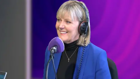 Joy Allen sitting in a BBC studio with headphones on and in front of a microphone. She is smiling and looking to her right side. She is wearing blue jacket and black polo top and necklace. The background is two different shades of purple.