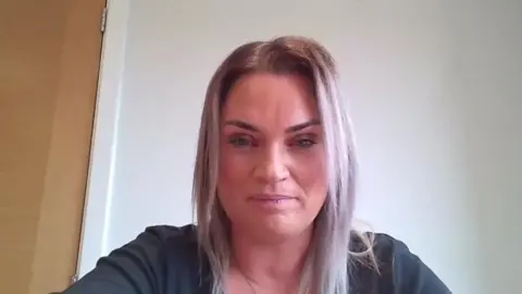 A woman called Nicola Hart sits down in front of a white wall and a wooden door. She has long straight hair going past her shoulders which looks like it has been dyed silver. She is wearing a black top. She has thick black eye-shadow on her eyebrows.