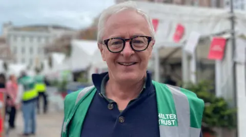 BBC A wide shot of Mike Powell. He has short grey hair and is wearing a dark green and grey Shelter Trust high visibility jacket.