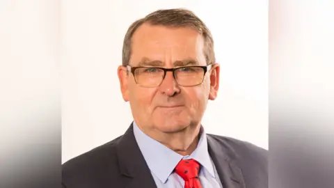 Stevenage Borough Council Headshot of Richard Henry with short dark hair, wearing a dark grey jacket, a light grey short and a red tie.