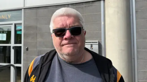 A man with short, grey hair and sunglasses smiles. He is wearing a black and amber Hull City jacket and is standing outside a football stadium.