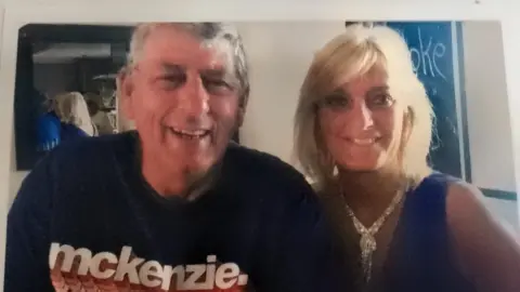 Marion Heard A man and a woman sitting close together indoors, smiling at the camera. The man is wearing a dark T‑shirt, and the woman wears a blue sleeveless top and a sparkly necklace.