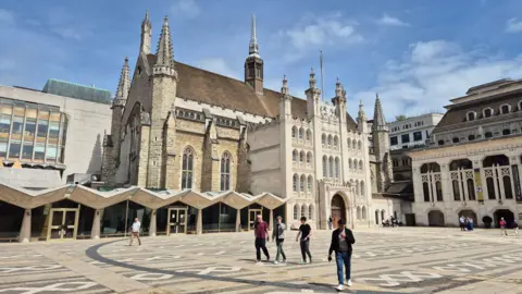 LDRS Image of Guildhall, which is home to the City of London Corporation. There are people walking around the area, which is surrounded by tall, old-fashioned, cathedral-like buildings. The concrete floor is covered in patterns. 