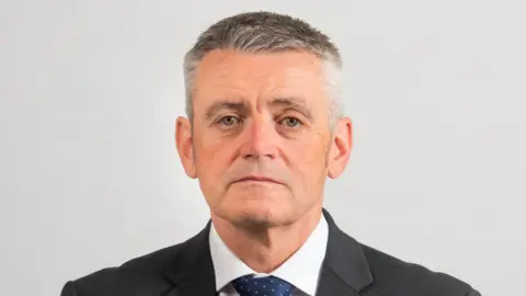 HMICFRS A man with short grey hair looks into the camera wearing a black suit with white shirt and navy tie. He sits in front of a white background.