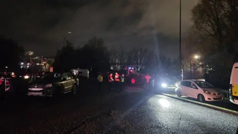 Mike Fennell A protest on a road. It is dark, but a tractor, many vehicles and some union jack flags can be seen. There are some people and police officers in high vis jackets standing on the road.