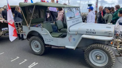 A light blue old open-topped military vehicle, with green seats and a green roof and big wheels and a flag on the back stands in front of a crowd of people.