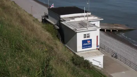 Geograph/Hugh Venables Sheringham lifeboat building in white with a navy lifeboat sign. A shingle beach directly below leads to the sea.