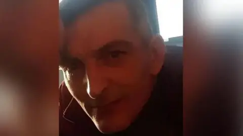 Police handout Paul Foster is photographed with brown hair and wearing a red jacket. There is a window behind him, letting in daylight.