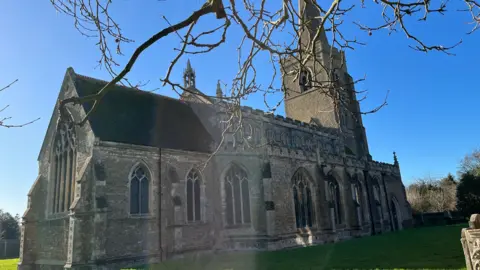 John Devine/BBC The outside of St Wendreda's Church in March. There is a blue sky and the spire can be seen at the far end of the building, with tree branches in the foreground.