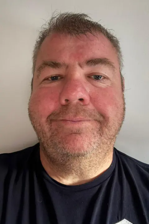 Sean O'Sullivan A man looking at the camera. He has a black t-shirt on and short hair. He has stubble