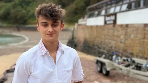 Cameron is wearing a white short-sleeved shirt stands on a brick slipway beside a small beach and harbour. Boats are moored in the water to the left, while a stone seawall and a waterfront building line the right side. Green hills rise in the background under an overcast sky.