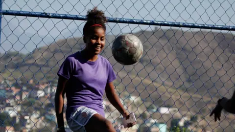 Gustavo Oliveira/WBR Photo Thaissa controls the ball with a solo kick, in front of a chain-link fence through which the grassy hills beyond the favela can be seen