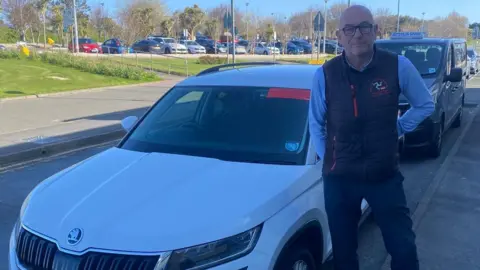 Mike Birchall, a bald man with glasses, he has a blue jumper and a gilet with the three legs of man, he smiles and stands next to his white taxi, at the rank outside of the airport.