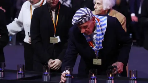 Getty Images Auschwitz survivors light candles as they pay tribute to Holocaust victims at a ceremony commemorating the 80th anniversary of the death camp's liberation. In the picture, an elderly man wearing a blazer and striped hat and scarf - like the stripes worn by prisoners - is lighting a candle.