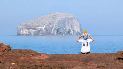 A man wearing a football shirt and with his child sitting on his shoulders looks across at an island on a clear, sunny day. 
