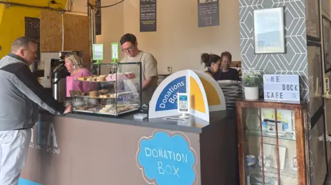 A man in grey jeans and a grey top is being served by a woman in pink with blond hair and a man with a cream tshirt on and glasses with short brown hair. There is a donations box sign in blue and yellow and some buns on show.