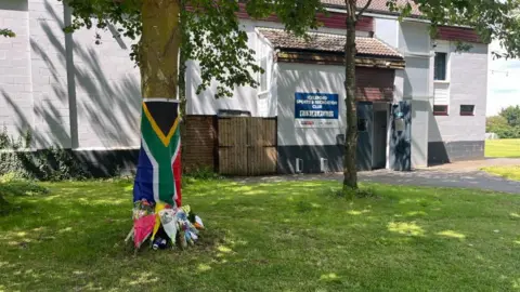 A tree trunk wrapped in a blue, green and red flag with bunches of flowers and the base. Behind is a wall and a sign saying "ICKLEFORD SPORTS & RECREATION CLUB"