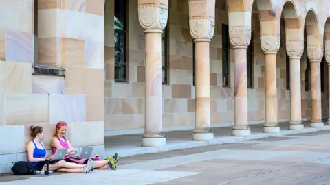 Two students sitting on the ground with laptops in front of a large building.