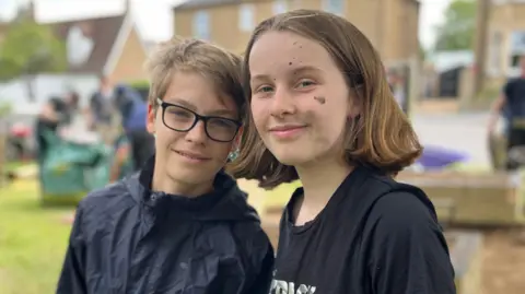 Daniel and Lyra are standing next to each other on a small patch of grass which is surrounded by houses. Daniel is standing on the left and has short brown hair, and is wearing black-framed glasses and a navy waterproof coat. Lyra is standing next to him on the right and has short brown hair, and is wearing a black T-shirt.