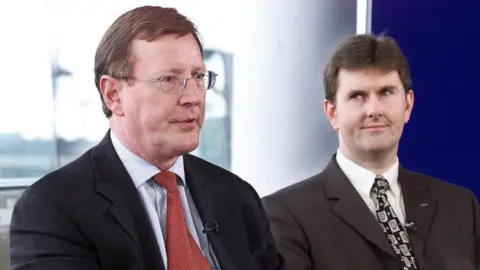 Getty Images An older photograph of two men. The older man on the left has short brown hair and is wearing small metal framed glasses. He is wearing a dark suit, light blue shirt and red patterned tie. The younger man on his left is looking at the older man with a raised eyebrow and slight grin. He he has black hair and is wearing a dark suit, white shirt and black patterned tie.