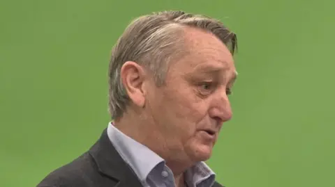 Anthony Slaughter holding his hands together in front of a microphone and a green backdrop, with the letter H visible.