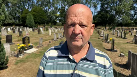 Phillip Norton/BBC A man in a blue and white striped polo shirt looks into the camera. He is standing in a cemetery. Rows of gravestones are visible in the background along with large green trees and shrubs.