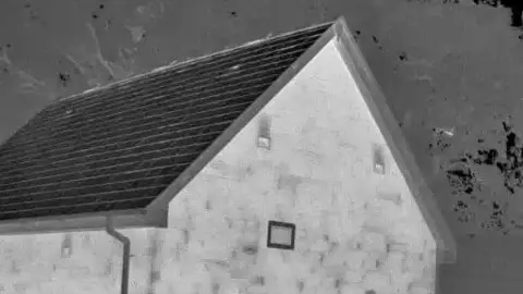 CEC A black and white grainy thermal image of a stone building with three small windows and a slate roof and a speck of bright light to the right which is the bat flying out