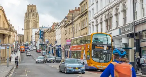 Park Street. It is a busy, hilled road with cars, busses and a cyclist travelling on it. The Wills Memorial building is at the top of it with a grand tower. Shops line the street and pedestrians walk up and down the pavement.
